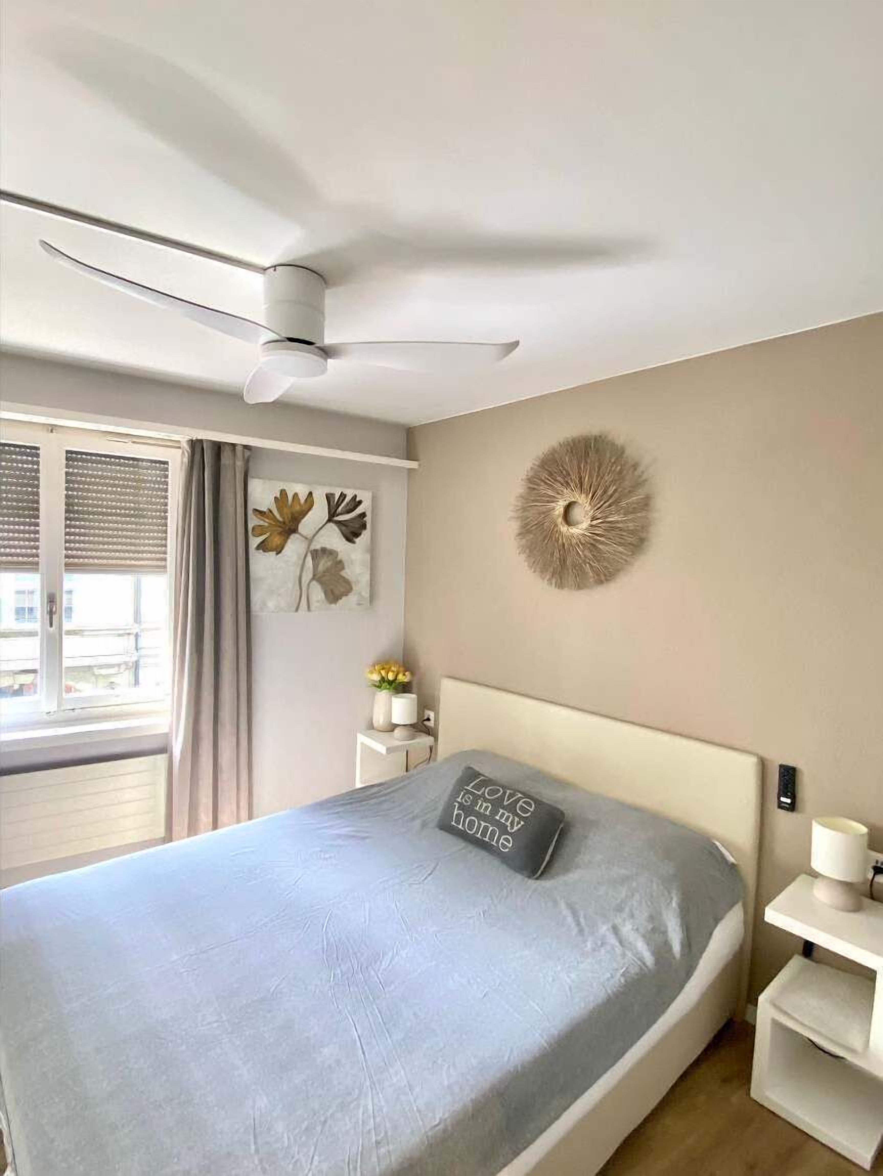 Een slaapkamer met een bed, een plafondventilator, een wanddecoratie, een nachtkastje met lamp en een raam met gordijnen.