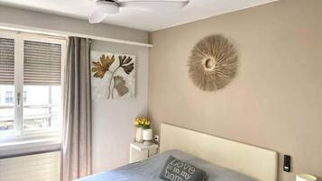 Een slaapkamer met een bed, een plafondventilator, een wanddecoratie, een nachtkastje met lamp en een raam met gordijnen.