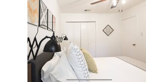 A bedroom with a bed, a desk lamp, a wall-mounted shelf, and a door with a decorative panel. - Woodstone Getaway (Austin)