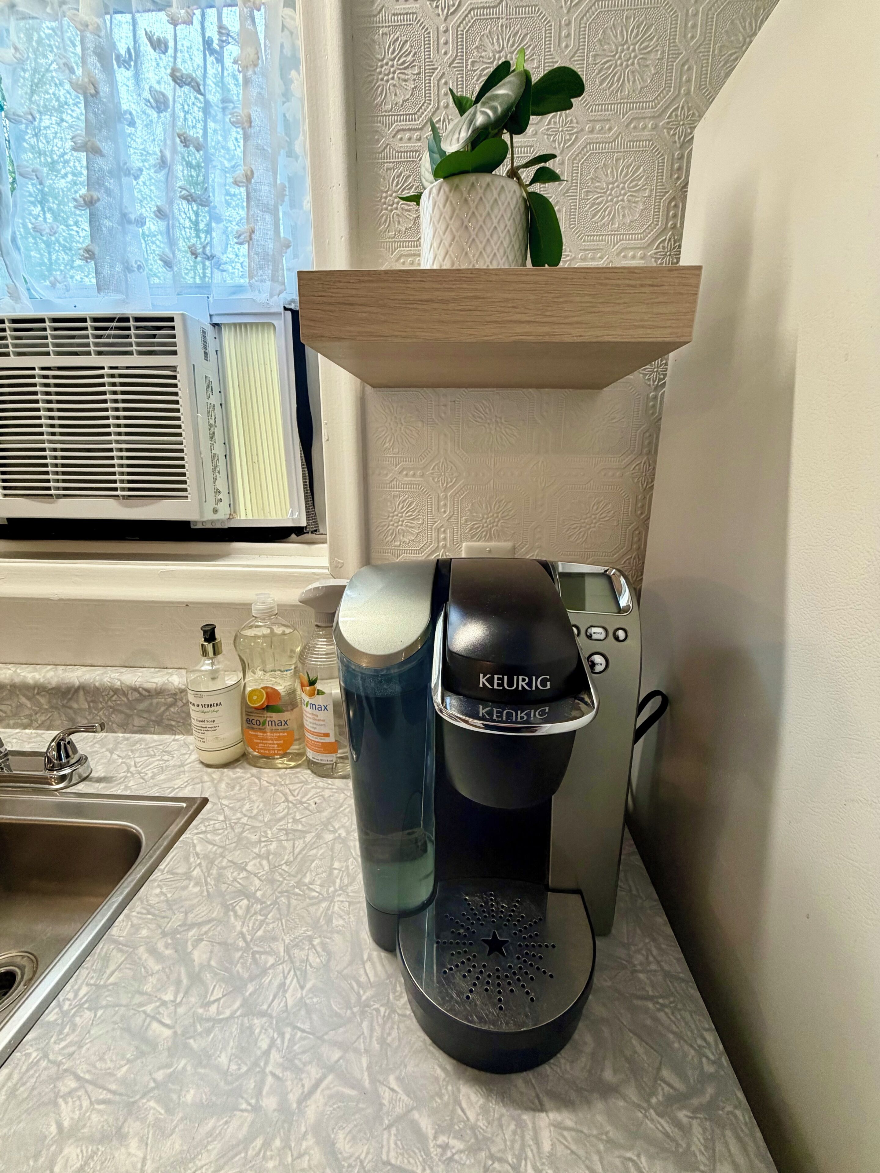 Eine Küchenzeile mit einer Keurig-Kaffeemaschine, einem Spülbecken und einem Fenster mit Klimaanlage.