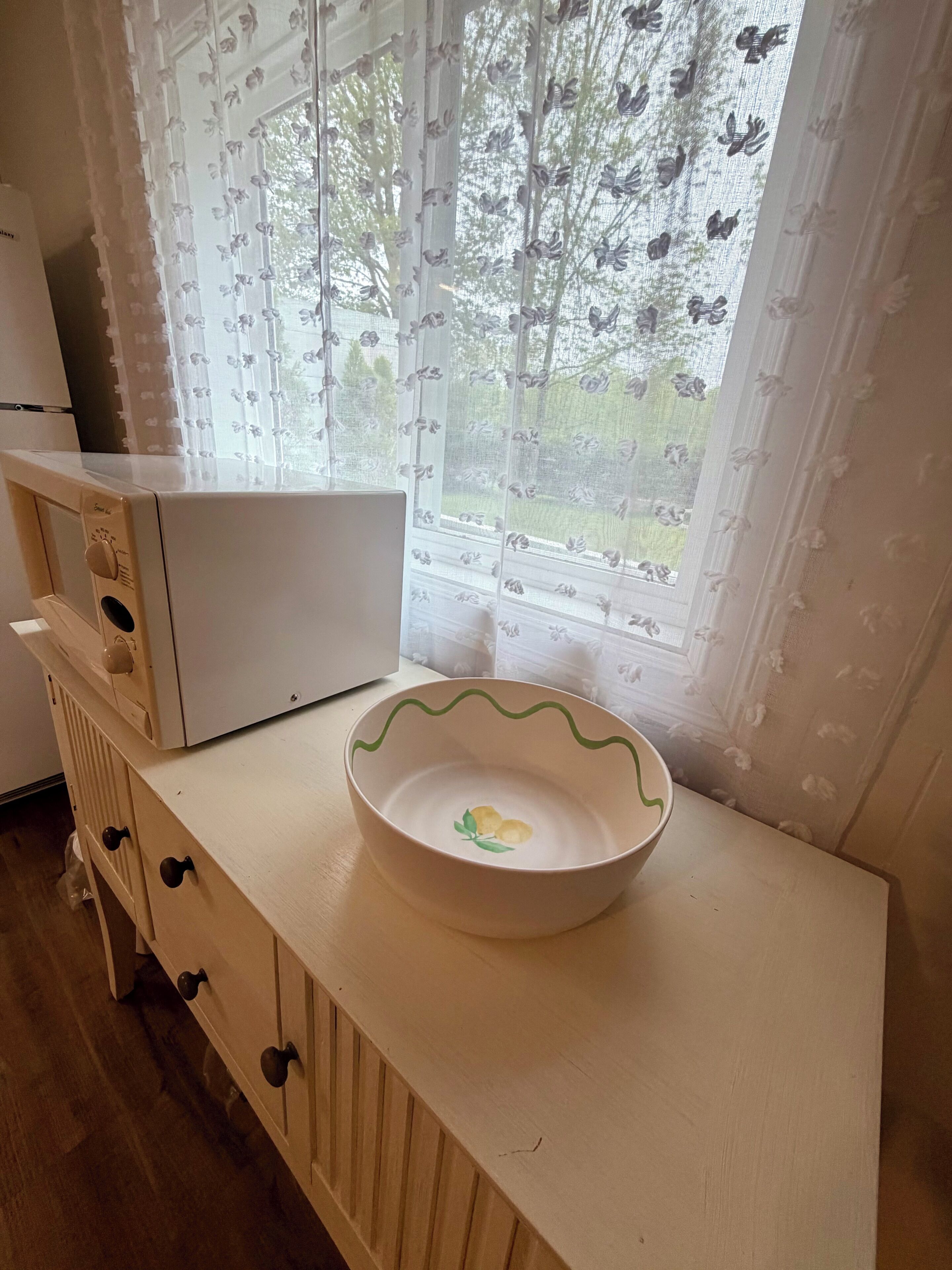 Ett kök med en mikrovågsugn, ett träskåp med lådor, en skål på bänkskivan och ett fönster med blommiga gardiner.