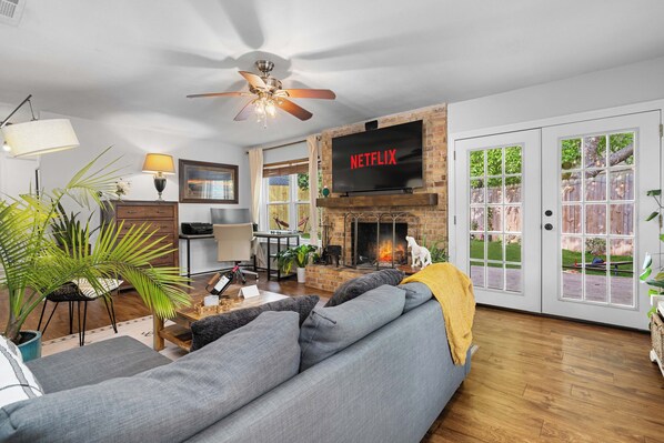 A living room with a fireplace, a sofa, a TV mounted above the fireplace, and a ceiling fan. - Woodstone Casita~Lush backyard~Central location (Austin)