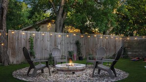 A backyard with a circular stone fire pit surrounded by four Adirondack chairs, a wooden fence, and string lights.