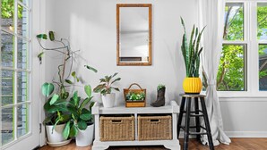 A room with a white console table, wicker baskets, potted plants, a mirror, and a window with a view of greenery.