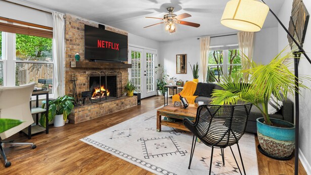 A living room with a fireplace, a TV mounted above it, a ceiling fan, and a large window with curtains.