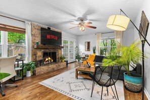 A living room with a fireplace, a TV mounted above it, a ceiling fan, and a large window with curtains. - Woodstone Casita~Lush backyard~Central location (Austin)