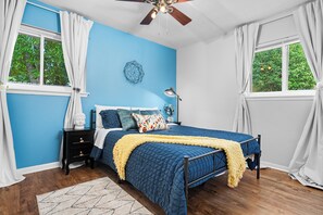 A bedroom with a blue wall, a bed with a blue quilt, a wooden floor, a ceiling fan, and a window with white curtains. - Woodstone Casita~Lush backyard~Central location (Austin)