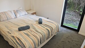 A bedroom with a bed, a nightstand, a drawer, and a window with a view of a forest. - 75 metres from small safe beach.  Sunsets to die for    (Auckland)