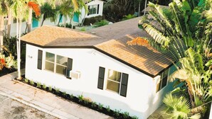 A white single-story house with a sloped roof, surrounded by palm trees and other greenery. - Villa #8 Pompano Beach: Spacious 1BR Canal View 4 Min Walk to Beach (Pompano Beach)