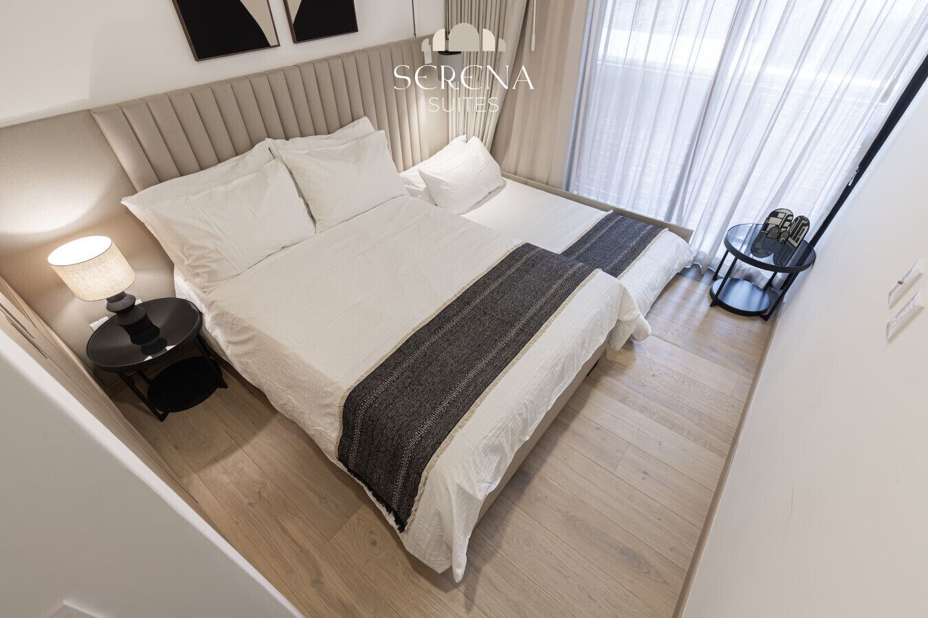 Une chambre d’hôtel comprenant un lit, des tables de chevet, une lampe, une petite table avec une chaise et une fenêtre avec des rideaux.