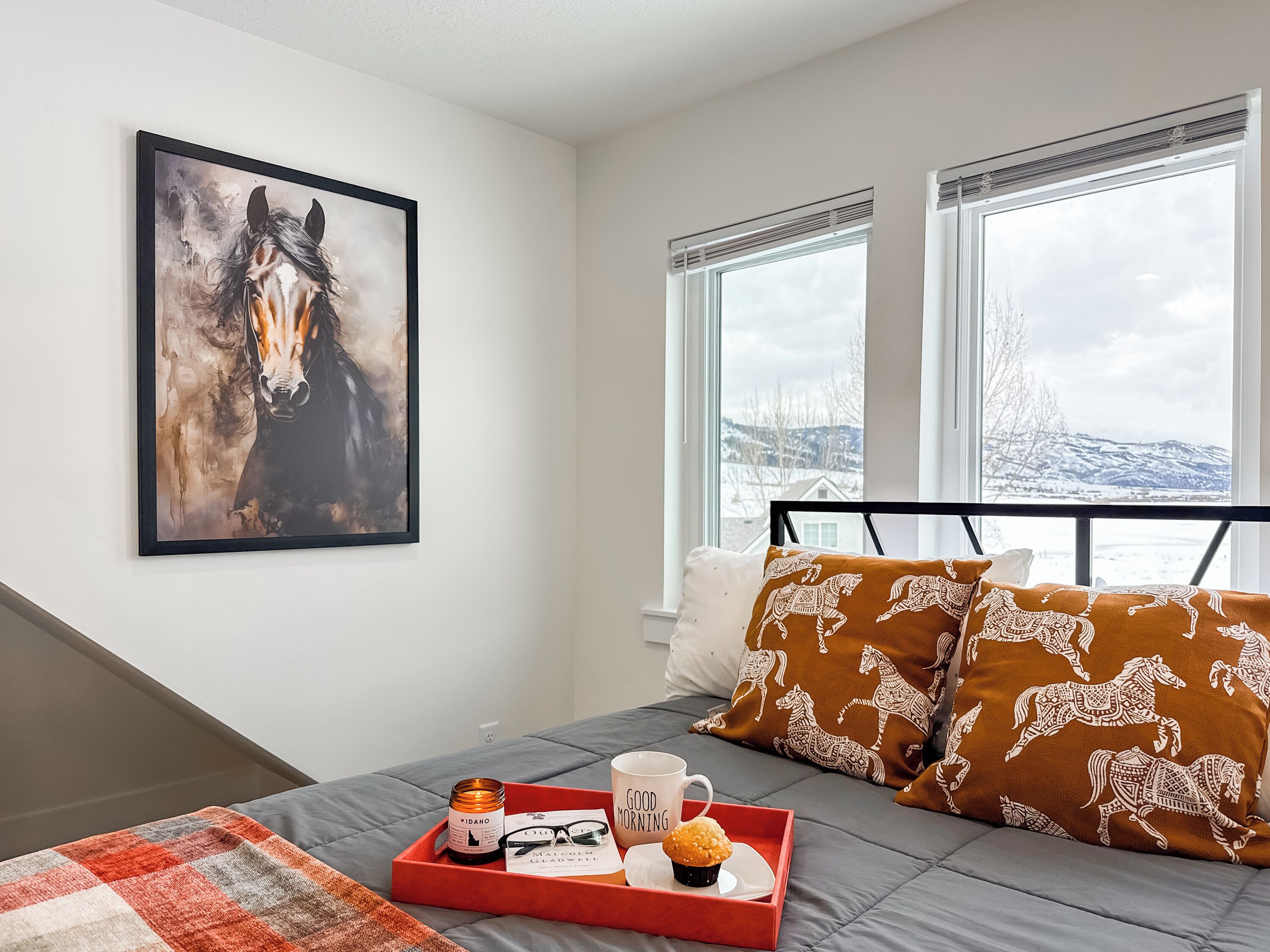 A bedroom with a bed, a framed horse painting, a window with a snowy view, and a tray with a candle, mug, and muffin.