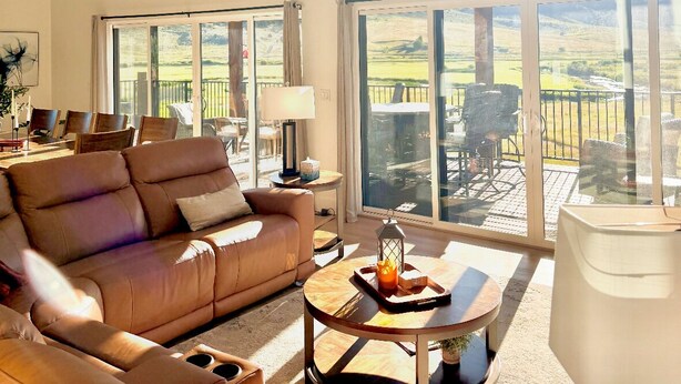 A spacious living room with a high ceiling, large windows, and a view of the outdoors. - Riverside Luxury: Private River Float with Hot Tub and 3 King Suites! (Lava Hot Springs)