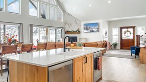 A spacious kitchen with a large island, wooden cabinets, and a view of a snowy landscape through large windows. - Riverside Luxury: Private River Float with Hot Tub and 3 King Suites! (Lava Hot Springs)