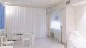 Een moderne, minimalistisch ingerichte kamer met een glazen tafel en witte stoelen, een grote schuifpui en een wit bed.