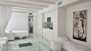 Een moderne hotelkamer met een glazen salontafel, een bed met witte beddengoed, een bank, een schilderij aan de muur en een keukenruimte zichtbaar achter een glazen wand.
