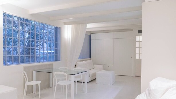 Een moderne, witte kamer met een glazen tafel en stoelen, een witte bank en een groot raam met jaloezieën.