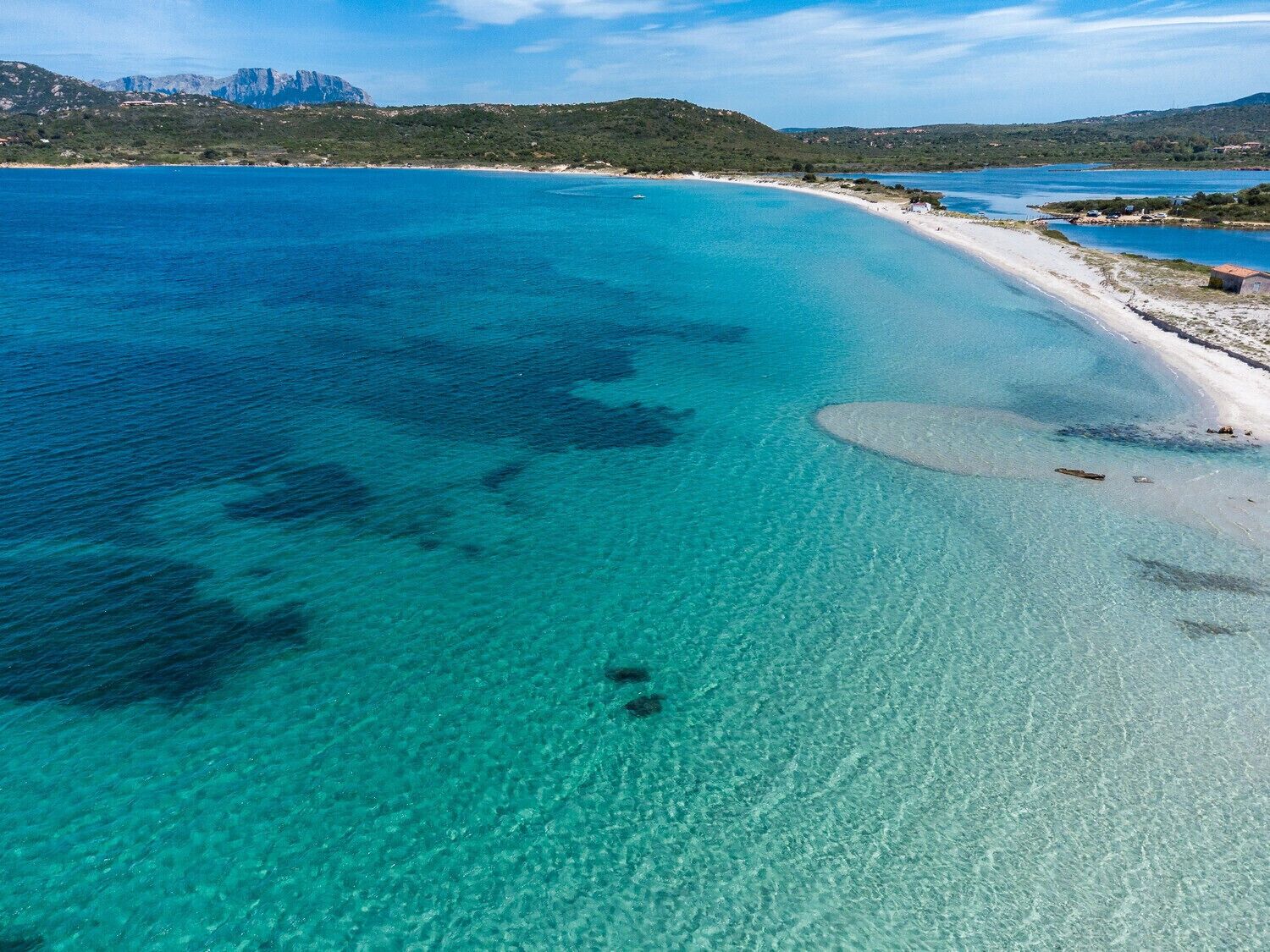 Eine Küstenlandschaft mit klarem türkisblauem Wasser, einem Sandstrand und einer hügeligen Landschaft im Hintergrund.