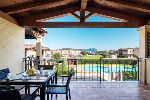 A covered outdoor area with a table and chairs, a view of a swimming pool, and residential buildings in the background.