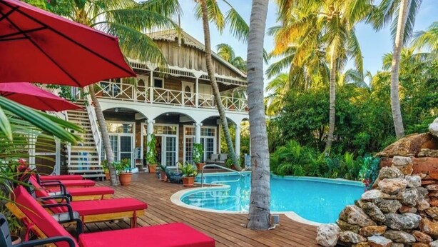 Une terrasse en bois avec des chaises longues rouges, une piscine et un bâtiment à toit de chaume, le tout entouré de palmiers.