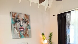 Une chambre à coucher avec un lit, un ventilateur de plafond, un tableau, une table de chevet avec une lampe et une fenêtre avec des rideaux.