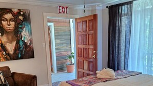Une chambre d’hôtel avec un lit, une chaise, un tableau au mur, un panneau de signalisation indiquant la sortie et un ventilateur de plafond.
