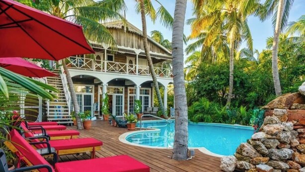 Un espace aménagé au bord de la piscine, avec des chaises longues rouges, une terrasse en bois et une piscine entourée de palmiers.