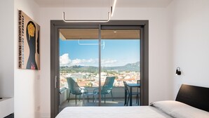 Quarto com cama grande, varanda com cadeiras e vista para a cidade.