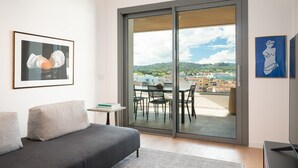 Uma sala de estar moderna com um sofá cinzento, uma mesinha com livros, uma varanda com mesa e cadeiras, e vista para a cidade.