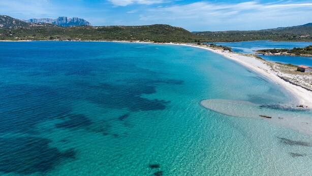 Vista aérea de uma área costeira com águas turquesa cristalinas, uma praia de areia e colinas verdes ao fundo.