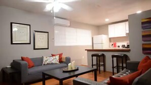 A modern living room with a grey sofa, a coffee table, and a ceiling fan. There is a kitchen area with white cabinets and a bar stool. - Midtown Gem! Sleeps 3 Near Richmond and Montrose (Houston)