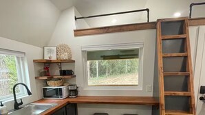 Cocina con alacenas de madera oscura, pileta de acero inoxidable, microondas y ventana con vista a árboles. Se observa una escalera de madera y dos taburetes.