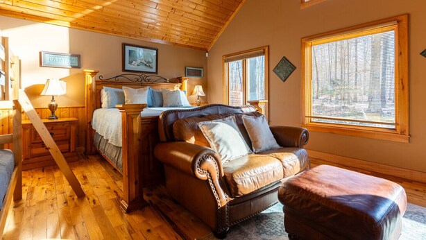 Una habitación acogedora con techo de madera, una cama con cabecera, un sofá de cuero y una ventana con persianas.
