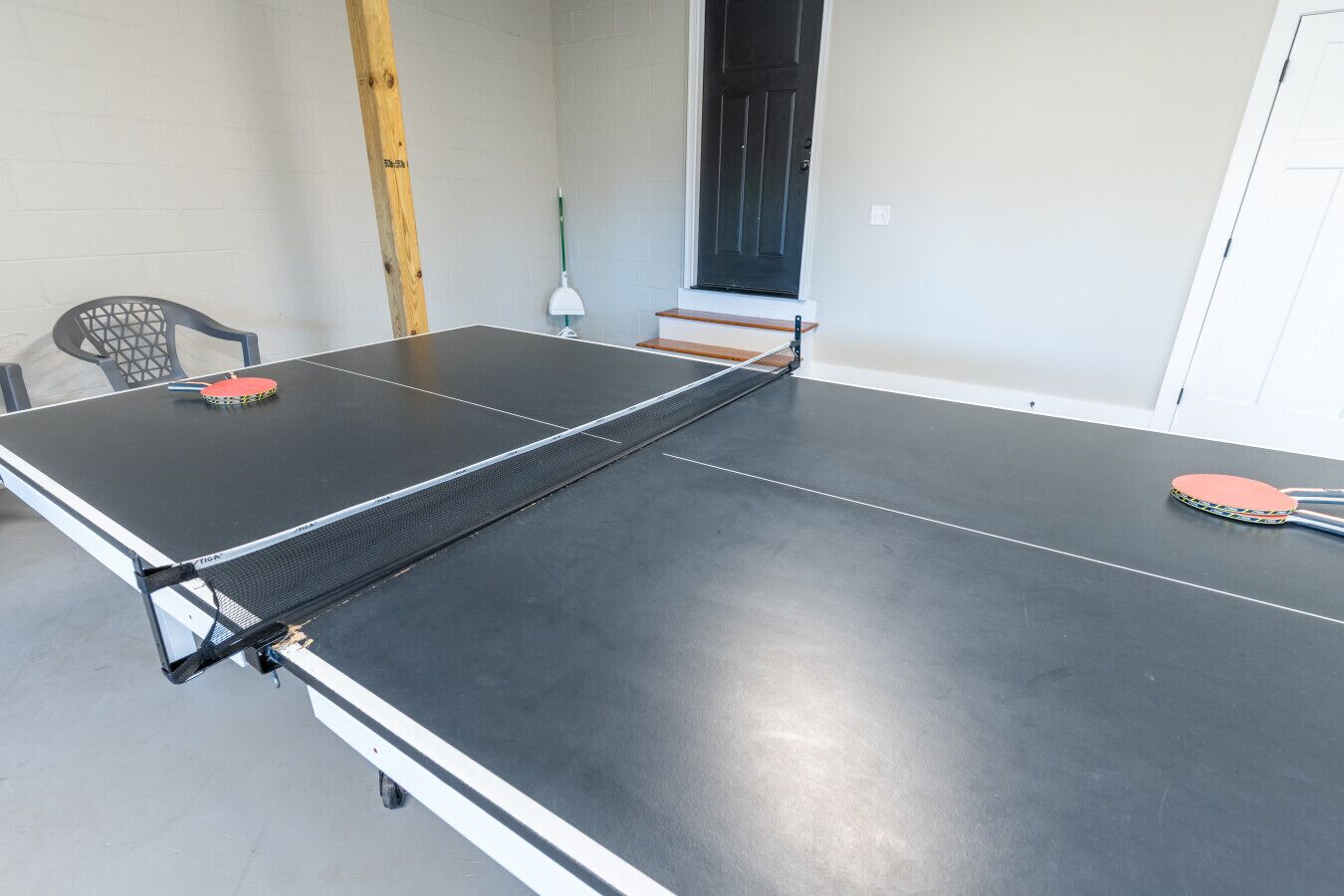 Tafeltennistafel met twee spatels en een bal, een stoel en een dweil in een kamer met witte muren en een deur.