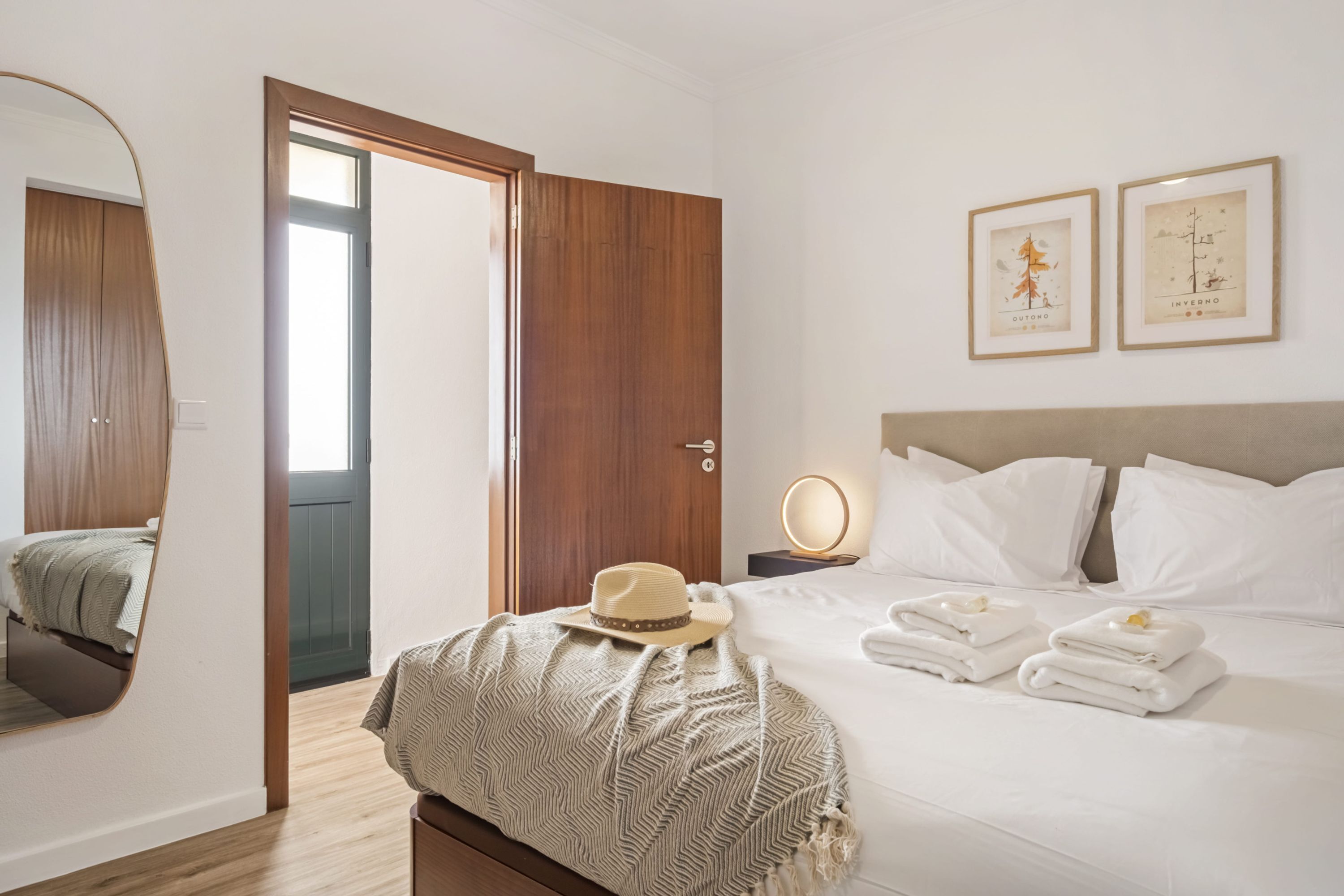A hotel room with a bed, a bedside table with a lamp, a mirror, a door, and a wall with framed pictures.