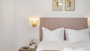Un letto rifatto con lenzuola bianche e una testiera rosa, un comodino con una lampada e un vaso di fiori, e due quadri appesi al muro.
