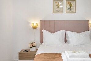 Una cama bien hecha con ropa de cama blanca y un cabecero rosa, una mesita de noche con una lámpara y un florero, y dos cuadros en la pared.