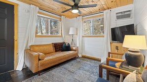 L’intérieur d’un chalet accueillant, avec un plafond en bois, un canapé en cuir, un tapis, une lampe et un téléviseur posé sur un meuble en bois.
