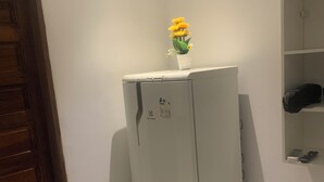 Un refrigerador blanco y compacto en un rincón de una habitación con paredes blancas, una puerta de madera y un estante con un control remoto.