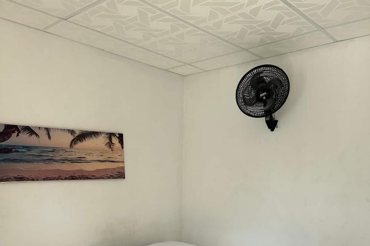 A single bed with a wooden frame and white bedding in a room with a beach-themed wall art and a wall-mounted fan.