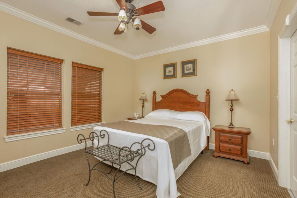 A bedroom with a bed, two bedside tables, a ceiling fan, and two windows with blinds.