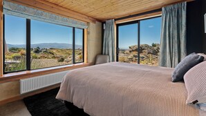 Un dormitorio con una cama grande, dos ventanas con cortinas y vistas a un paisaje rocoso, además de un techo de madera.