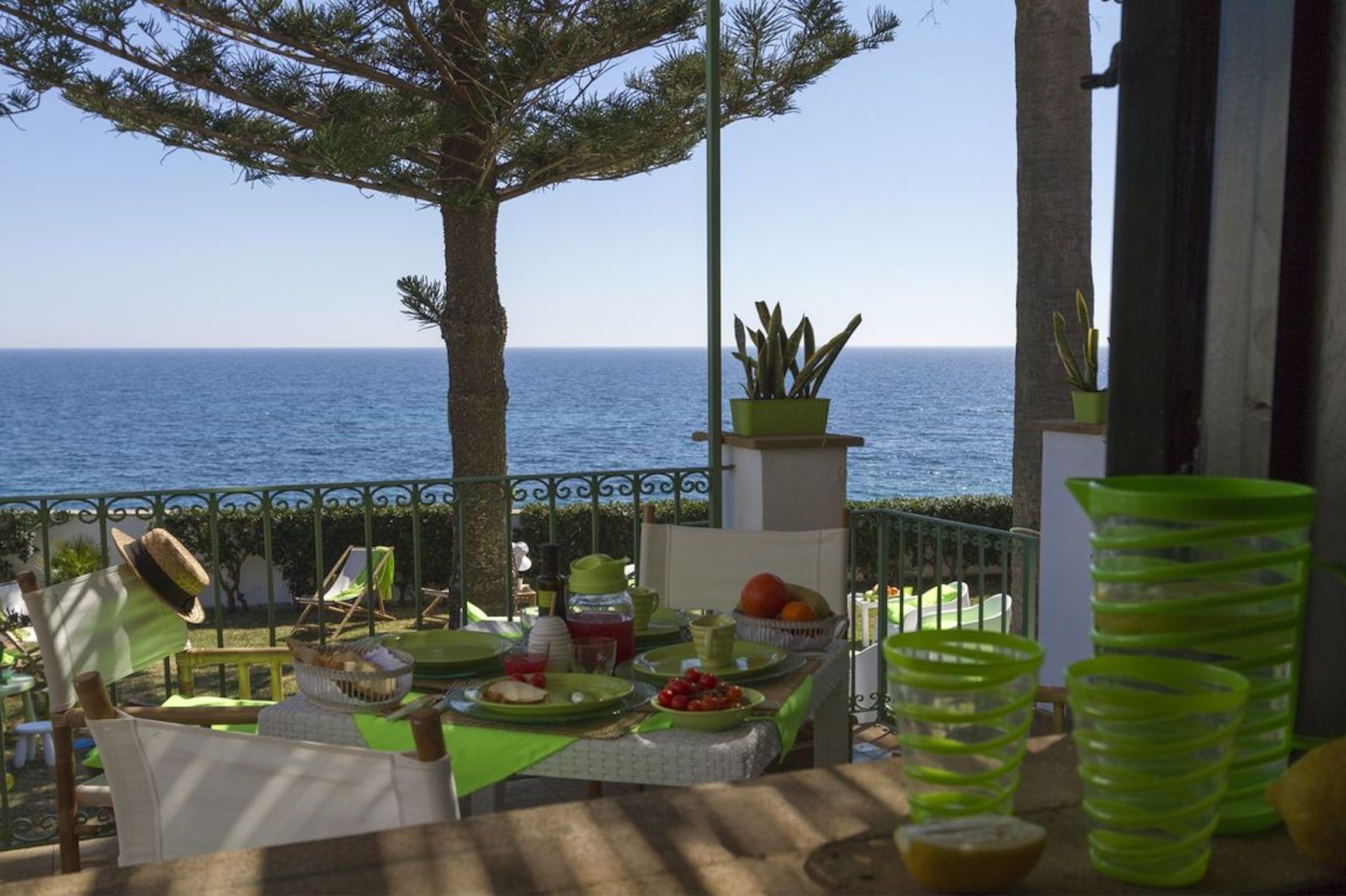 A terrace with a table set for a meal, overlooking the sea, with potted plants and outdoor furniture.