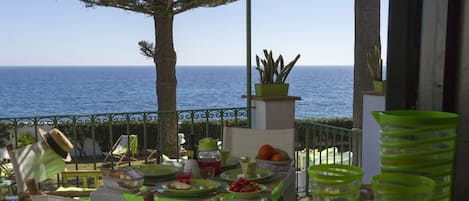 A terrace with a table set for a meal, overlooking the sea, with potted plants and outdoor furniture.