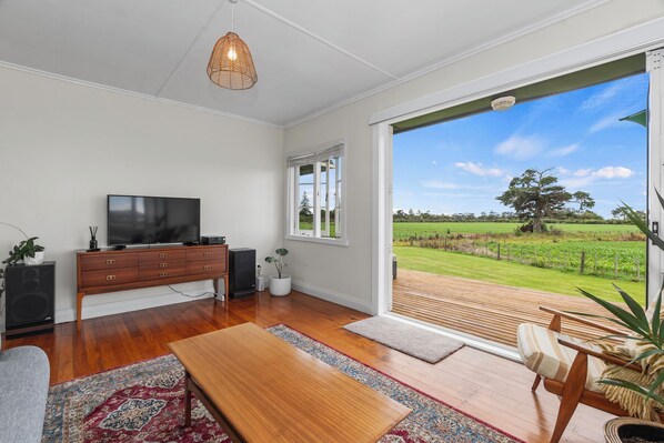 Een ruime woonkamer met een grote televisie, houten meubels en uitzicht op een groen veld door een schuifglazen deur.
