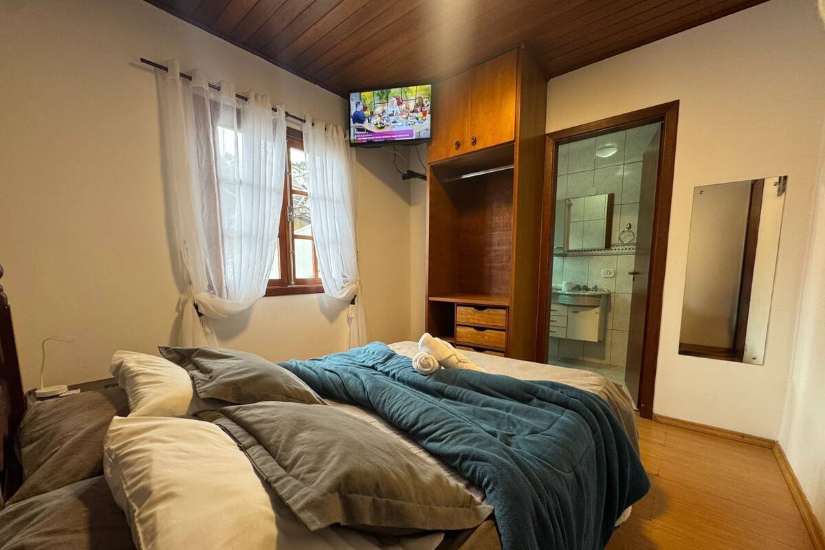 A bedroom with a bed, a TV mounted on the wall, a window with curtains, a wooden wardrobe, and a bathroom with a mirror.