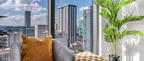 A modern living room with a grey armchair, a patterned ottoman, a potted plant, and a large window offering a city view.