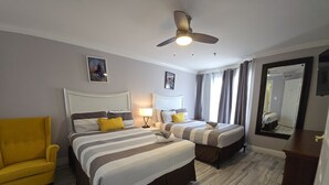 Una habitación de hotel con dos camas, un sillón amarillo, un ventilador de techo, un espejo y un televisor.