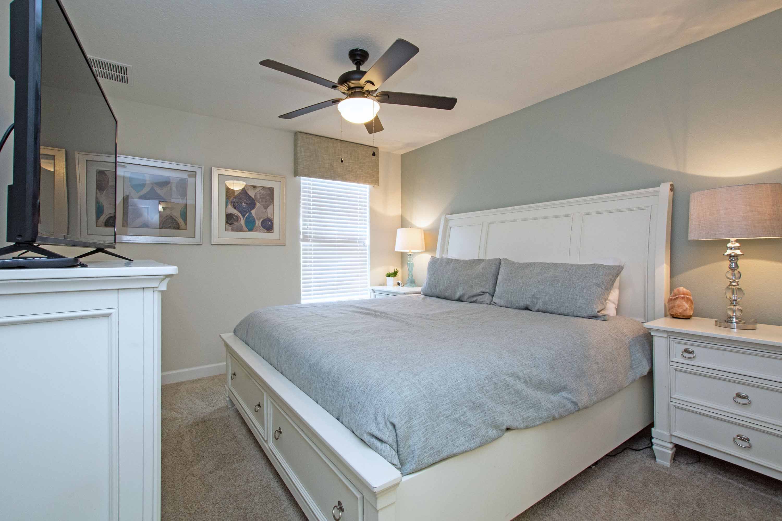 A bedroom with a bed, a nightstand, a lamp, a ceiling fan, and framed pictures on the wall.