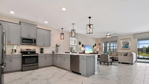 A modern kitchen with grey cabinets, stainless steel appliances, and a marble floor. There is a dining area with a sofa and a TV. - Home 9 Bed Pool Best Location Storey Lake (Kissimmee)