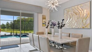 A dining area with a large table, cushioned chairs, a modern chandelier, and a view of a swimming pool and greenery. - Home 9 Bed Pool Best Location Storey Lake (Kissimmee)